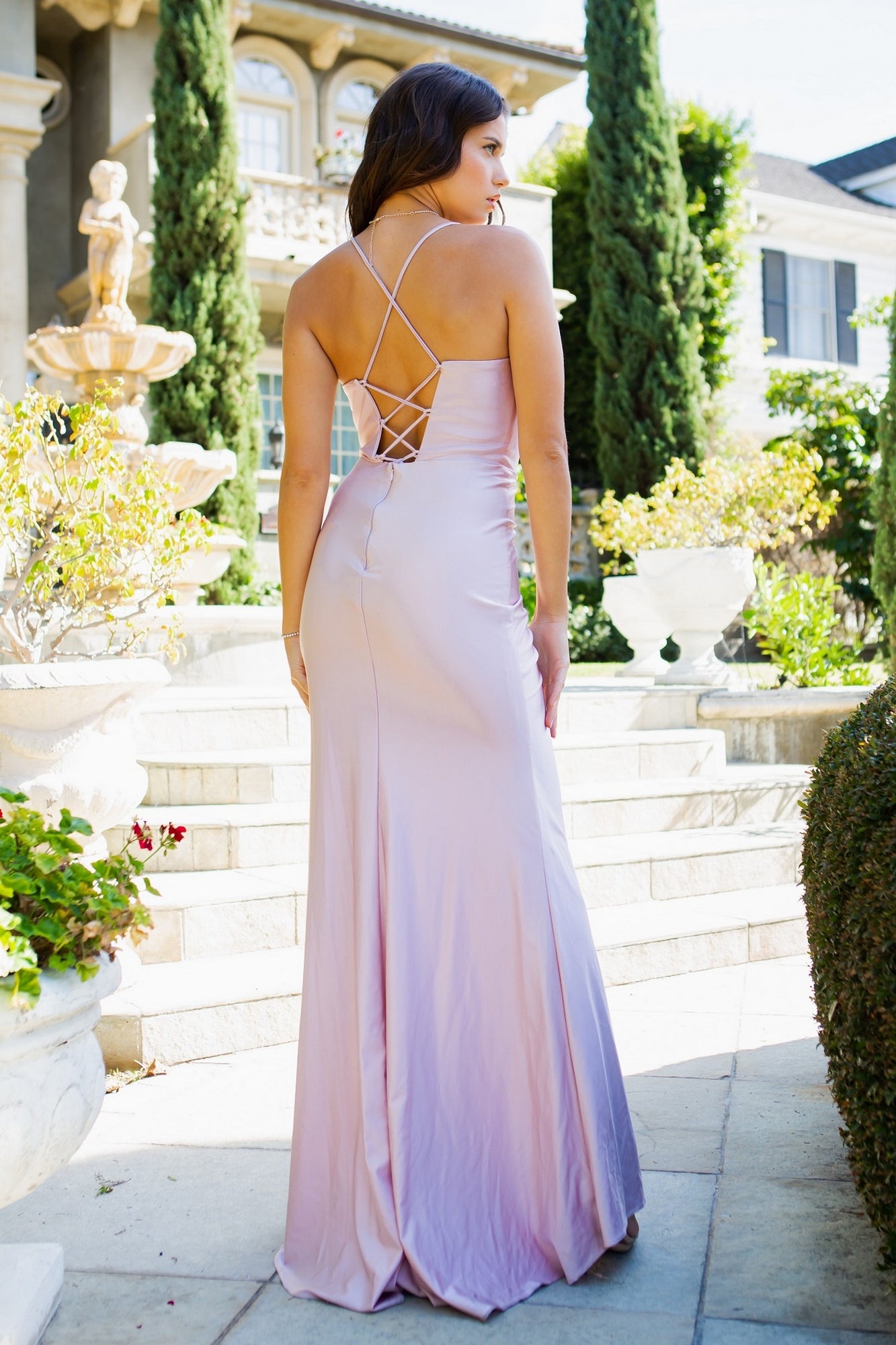A woman stands outdoors on stone steps, facing away, wearing the Cowl-Neck Long Formal Corset Prom Dress 8035J in light purple with crisscross open-back straps.
